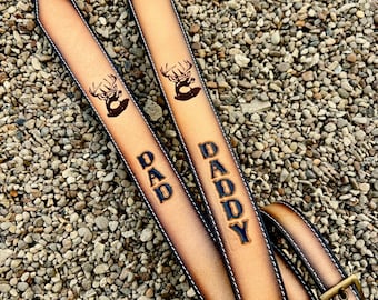 May include: Two leather guitar straps with the words "DAD" and "DADDY" embossed in black. Each strap features a deer head design and white stitching. The straps have brass-colored buckles and black end pieces.