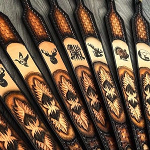 May include: A collection of brown leather gun slings with various wildlife designs, including deer, turkey, and a moose. The slings have a black leather trim and a woven pattern.