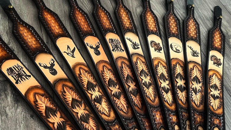 May include: A collection of brown leather gun slings with various wildlife designs, including deer, turkey, and a moose. The slings have a black leather trim and a woven pattern.