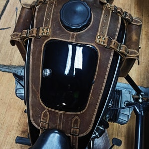 May include: A brown leather motorcycle gas tank cover with a black gas cap and a black leather seat. The cover has a flap with a buckle and a strap with a buckle.