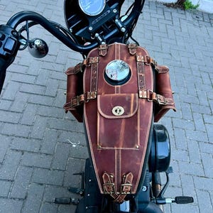 Könnte beinhalten: Braune Motorrad-Tanktasche aus Leder mit kleiner Tasche und dekorativen Schnallen. Die Tasche ist an einem schwarzen Motorrad montiert, mit einem Tachometer und Lenker im Hintergrund. Die Tasche hat einen runden Spiegel.