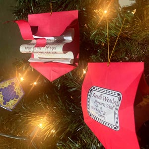 May include: Two red paper ornaments hanging on a Christmas tree. The top ornament has a rolled up scroll inside. The bottom ornament has a white label with black text that reads "Ronald Weasley Hogwarts School Witchcraft & Wizardry".
