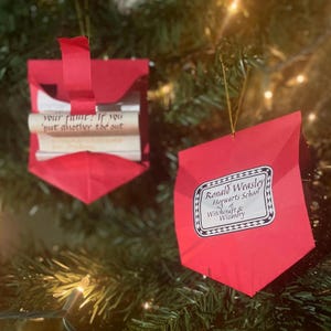May include: Two red paper ornaments shaped like envelopes with a gold string for hanging. One ornament has a rolled up scroll inside with the text "your fame? If you but another the out". The other ornament has a white label with black text that reads "Ronald Weasley Hogwarts School of Witchcraft & Wizardry".