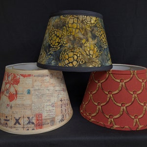 May include: Three lampshades with different patterns. The top lampshade has a dark blue background with a yellow and brown turtle pattern. The middle lampshade has a beige background with a red, white, and black pattern. The bottom lampshade has a red background with a gold pattern.