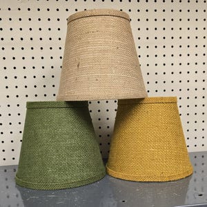 May include: Three conical lamp shades with a textured woven finish. One shade is light tan, one is olive green, and one is mustard yellow. Each shade features a simple, tapered design with a finished top and bottom edge, arranged in a stacked formation.