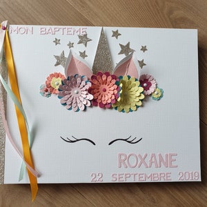 Peut inclure: Un album blanc à spirales avec un motif de licorne. La couverture présente une corne scintillante, des oreilles roses et des fleurs en papier colorées. Le texte "MON BAPTÊME" et "ROXANE 22 SEPTEMBRE 2019" sont affichés en rose.