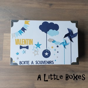 May include: A white keepsake box with a blue and yellow design. The box has the name "Valentin" written on it, along with a bow tie and stars. The text "Boite a Souvenirs" is written on the bottom of the box.