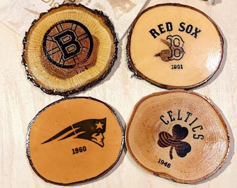 May include: Four wooden coasters with different sports team logos. The coasters are round and have a natural wood finish. The logos are black and white and include the Boston Bruins, Boston Red Sox, New England Patriots, and Boston Celtics.