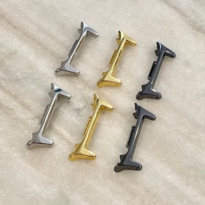 May include: Six small, metallic letter "I" charms in silver, gold, and black. The charms are arranged on a light-colored, marbled surface. The charms are likely for jewelry making or crafting.
