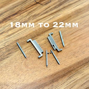 May include: A collection of silver-colored watch band spring bars and clasps on a wooden surface. The text "18MM TO 22MM" is displayed in white. The items are designed for watch band repair or replacement.