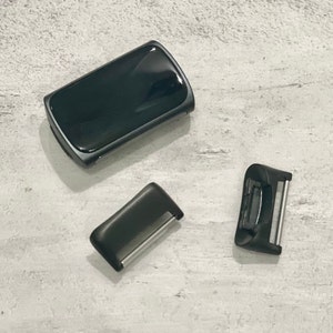 May include: Three black watch band components are displayed on a light gray surface. The components include a rectangular watch face, a small rectangular piece with a silver bar, and a buckle with a silver bar.