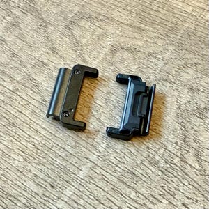 May include: Two black metal watch strap connectors. One has a hinged design, the other a sliding mechanism. Both are rectangular with rounded edges, designed to attach to a watch strap.