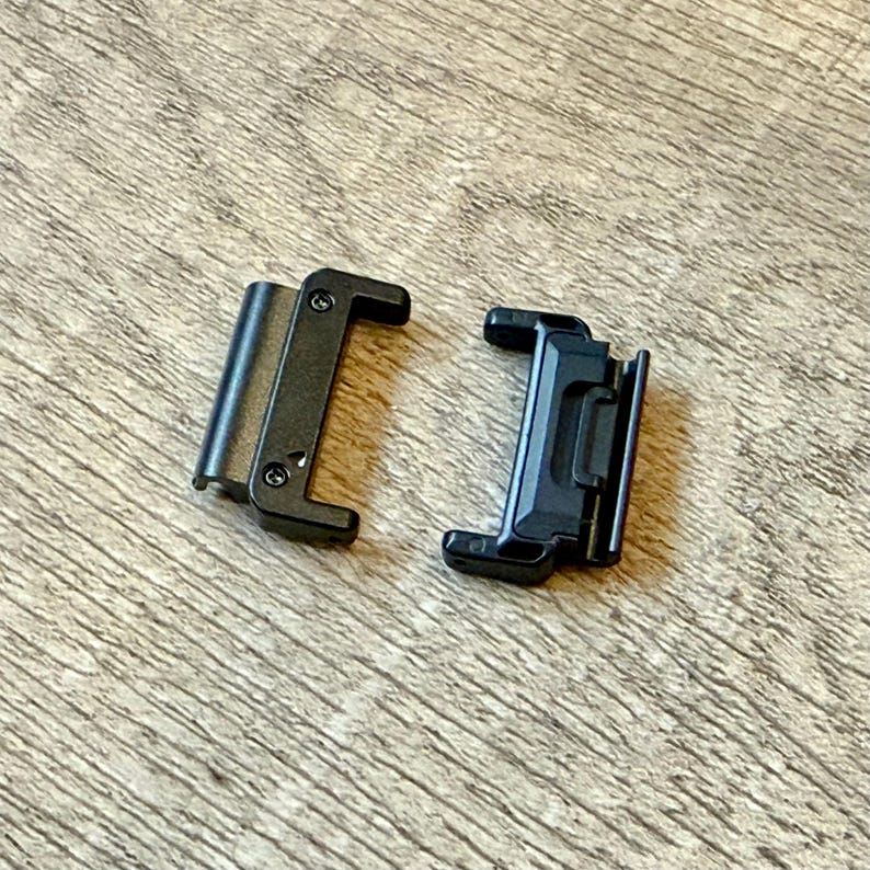 May include: Two black metal watch strap connectors. One has a hinged design, the other a sliding mechanism. Both are rectangular with rounded edges, designed to attach to a watch strap.