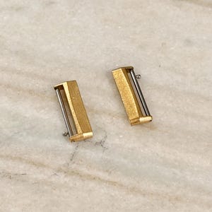 Puede incluir: Dos cierres de correa de reloj de metal dorado. Cada cierre tiene forma rectangular con acabado dorado cepillado y una barra plateada. Los cierres est&aacute;n colocados sobre una superficie clara, similar al m&aacute;rmol.
