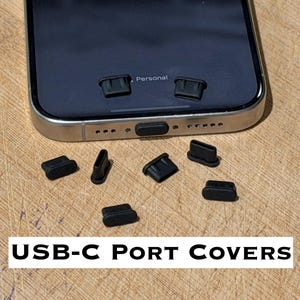 May include: Black USB-C port covers scattered on a wooden surface near a smartphone. The covers are small, rectangular, and designed to protect the phone's charging port. The image includes the text "USB-C PORT COVERS".