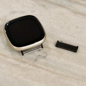 Puede incluir: Un reloj inteligente con pantalla negra y marco plateado. Un componente de correa de reloj negro est&aacute; separado y colocado junto a la esfera del reloj. El reloj est&aacute; sobre una superficie de m&aacute;rmol.