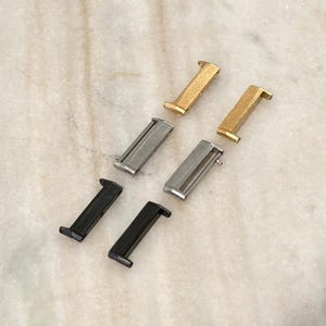 May include: A selection of metal clasps in gold, silver, and black. The clasps are rectangular with a hook or bar design, suitable for jewellery or craft projects. The clasps are arranged on a light-coloured, marble-like surface.