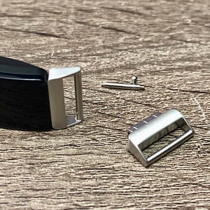May include: A black fitness tracker band with a silver metal clasp and a small silver pin. The band is lying on a wooden surface.