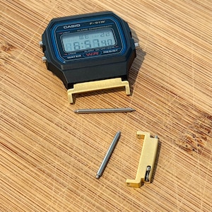 Puede incluir: Un reloj digital Casio F-91W negro con un borde azul alrededor de la pantalla, que muestra el texto CASIO, F-91W, ALARM CHRONOGRAPH y WATER RESIST. Se incluyen dos adaptadores de correa de metal dorado y dos pasadores de resorte plateados.