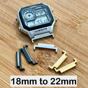 Könnte beinhalten: Eine silberne Casio Illuminator Digitaluhr mit quadratischem Zifferblatt, Weltkartenanzeige und Alarmeinstellungen. Unter der Uhr befinden sich drei Metall-Armbandadapter in Schwarz, Silber und Gold, beschriftet mit 18mm bis 22mm.