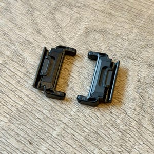 May include: Two black plastic clips with a rectangular design and a hinged section. The clips are positioned on a light brown wooden surface. Each clip has a series of small holes and a locking mechanism.