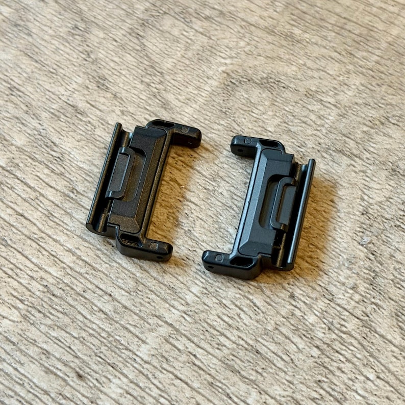 May include: Two black plastic clips with a rectangular design and a hinged section. The clips are positioned on a light brown wooden surface. Each clip has a series of small holes and a locking mechanism.