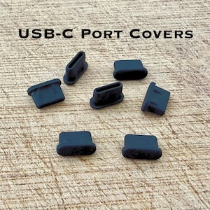 May include: A collection of dark blue USB-C port covers. The covers are in various orientations, some rectangular and some with an open slot. The text "USB-C PORT COVERS" is displayed in black at the top of the image.
