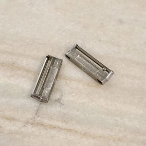 Puede incluir: Dos peque&ntilde;os cierres met&aacute;licos rectangulares con acabado plateado. Cada cierre tiene un marco rectangular con una barra deslizante y una placa central. Los cierres est&aacute;n colocados sobre una superficie clara, similar al m&aacute;rmol.