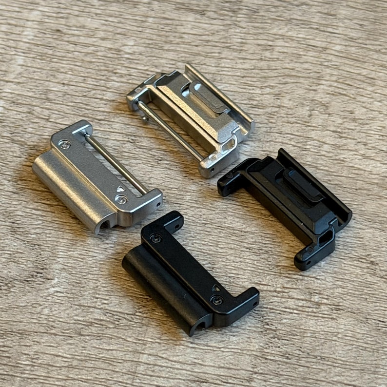 May include: Four metal watch strap clasps in silver and black. The clasps are rectangular with a hinged design and a metal bar. The clasps are arranged on a wooden surface.