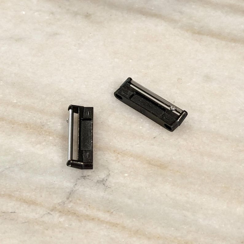 Puede incluir: Dos peque&ntilde;os cierres met&aacute;licos negros con detalles plateados. Un cierre est&aacute; en posici&oacute;n vertical, el otro en horizontal. Los cierres est&aacute;n sobre una superficie clara y veteada. Estos cierres probablemente se utilizan para asegurar objetos.