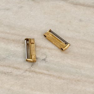 Puede incluir: Dos peque&ntilde;os cierres rectangulares de metal dorado con una barra plateada. Los cierres est&aacute;n sobre una superficie clara, similar al m&aacute;rmol. Los cierres parecen ser componentes o hallazgos de joyer&iacute;a.