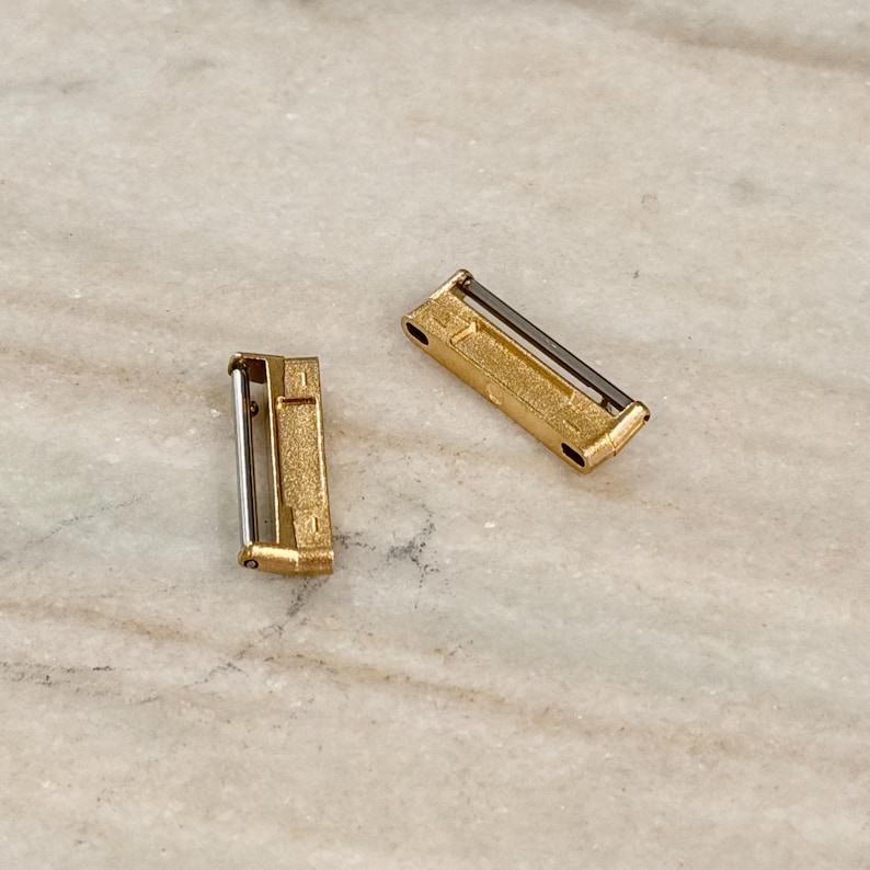 Puede incluir: Dos peque&ntilde;os cierres rectangulares de metal dorado con una barra plateada. Los cierres est&aacute;n sobre una superficie clara, similar al m&aacute;rmol. Los cierres parecen ser componentes o hallazgos de joyer&iacute;a.