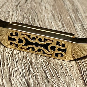 Puede incluir: Puente de guitarra de metal dorado con un intrincado diseño de filigrana negra.