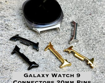 Puede incluir: Una caja de reloj Galaxy Watch 9 plateada con pantalla circular negra, acompañada de cinco conectores de correa de metal en acabados negro, dorado y plateado. El texto Galaxy Watch 9 Connectors 20mm Pins está impreso en la parte inferior.