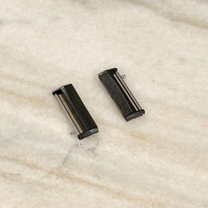 Puede incluir: Dos conectores de correa de reloj negros con detalles plateados. Cada conector tiene forma rectangular con un pasador plateado en un extremo. Los conectores se muestran sobre una superficie clara, similar al m&aacute;rmol.