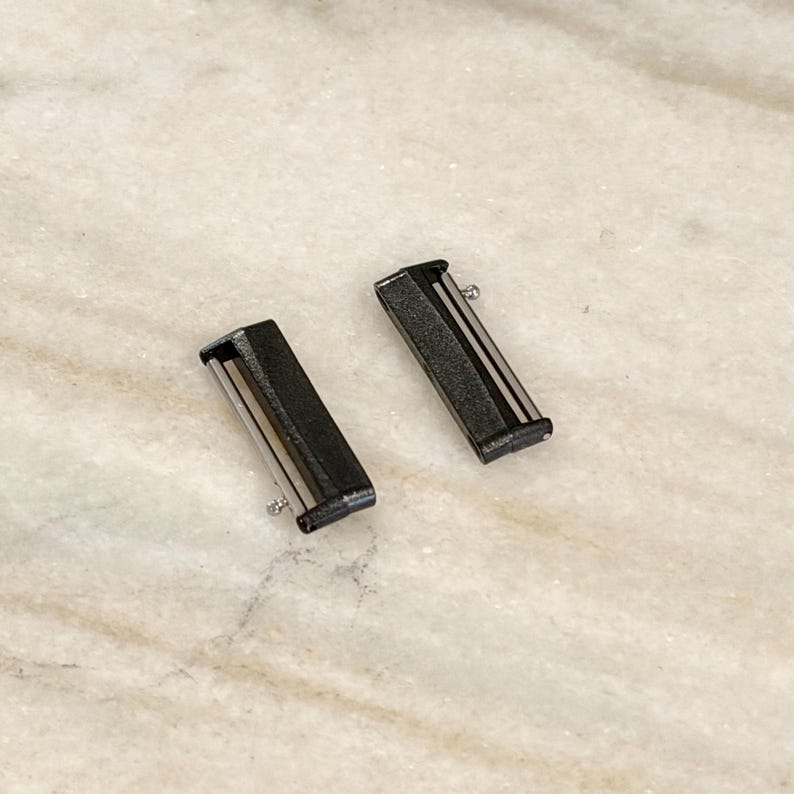 Puede incluir: Dos conectores de correa de reloj negros con detalles plateados. Cada conector tiene forma rectangular con un pasador plateado en un extremo. Los conectores se muestran sobre una superficie clara, similar al m&aacute;rmol.