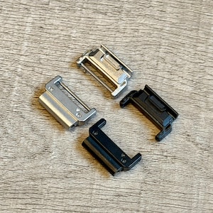 May include: Four metal watch strap clasps in silver and black are arranged on a wooden surface. The clasps have a rectangular shape with a hinged design and a metal bar. The clasps are likely replacement parts for a watch strap.