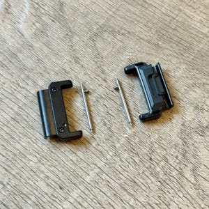 May include: Two black watch strap clasps and two silver spring bars are displayed on a wooden surface. The clasps are rectangular with rounded edges and have a matte finish. The spring bars are thin, cylindrical, and metallic.