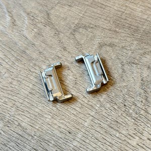 May include: Two silver metal watch clasps. Each clasp has a rectangular shape with a hinged section and a locking mechanism. The clasps are on a wooden surface with a natural grain pattern.