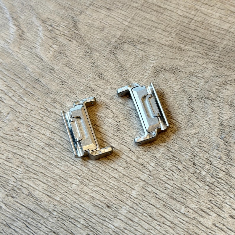 May include: Two silver metal watch clasps. Each clasp has a rectangular shape with a hinged section and a locking mechanism. The clasps are on a wooden surface with a natural grain pattern.