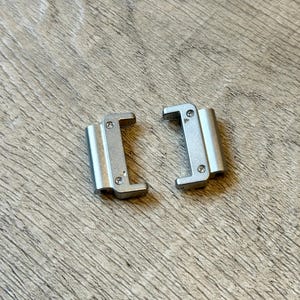 May include: Two silver metal watch strap connectors. Each connector has a rectangular body with a curved top and a small screw on each side. The connectors are on a light brown wooden surface.
