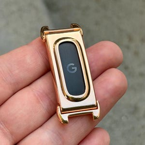 May include: A rose gold-toned metal watch accessory featuring a rectangular frame with rounded corners and a central black inlay. The center of the black inlay displays a small, light gray letter G logo. The frame has integrated attachment lugs.