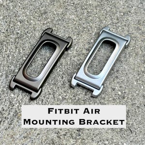 May include: Two rectangular Fitbit Air mounting brackets, one in a dark charcoal finish and one in a brushed silver finish. Each bracket features a central elongated oval cutout and integrated attachment points at the top and bottom ends.