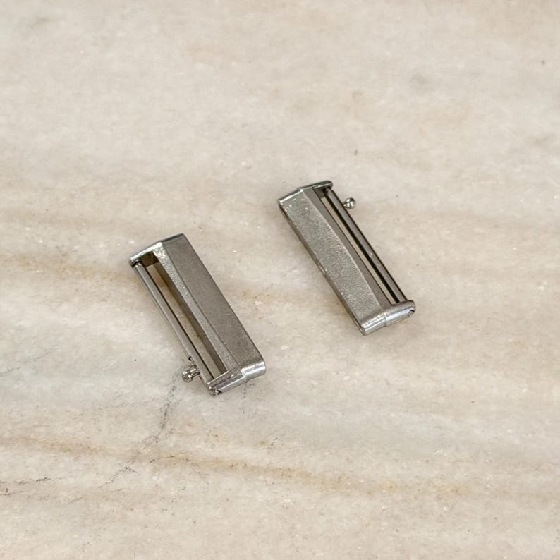 Puede incluir: Dos cierres plateados para correas de reloj. Cada cierre es rectangular con una barra deslizante y un peque&ntilde;o pasador. Los cierres est&aacute;n sobre una superficie clara y texturizada.