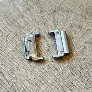 May include: Two silver metal watch strap connectors. Each connector has a rectangular shape with rounded edges and small screws. The connectors are designed to attach a watch strap to a watch case. The background is a light brown wooden surface.