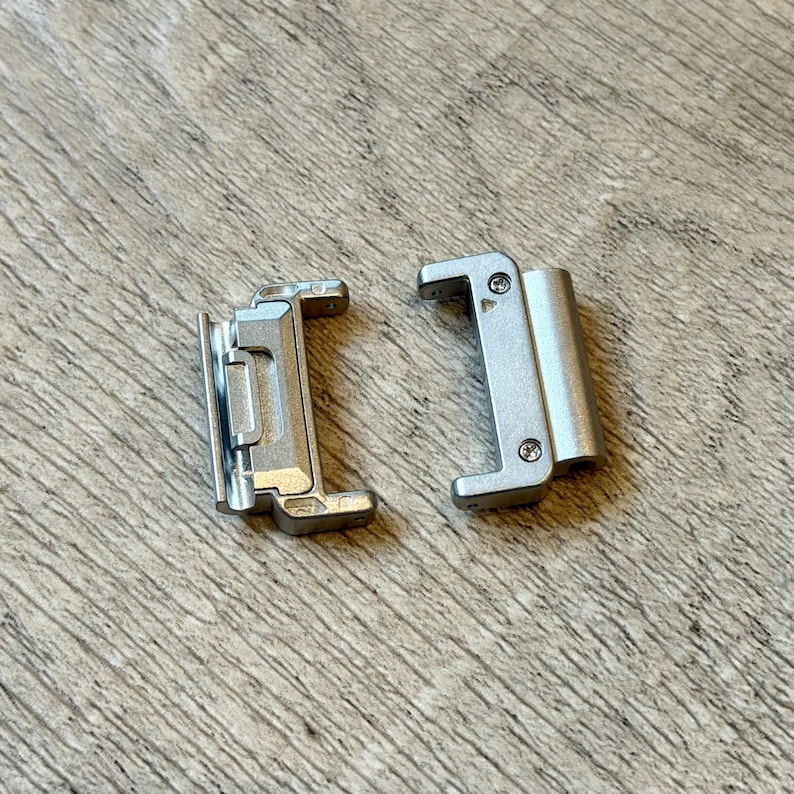May include: Two silver metal watch strap connectors. Each connector has a rectangular shape with rounded edges and small screws. The connectors are designed to attach a watch strap to a watch case. The background is a light brown wooden surface.