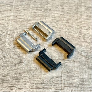 May include: Four watch strap clasps in silver and black. Two silver clasps are rectangular with rounded edges, and one has a silver pin. Two black clasps have a similar design. All are on a light wood grain surface.