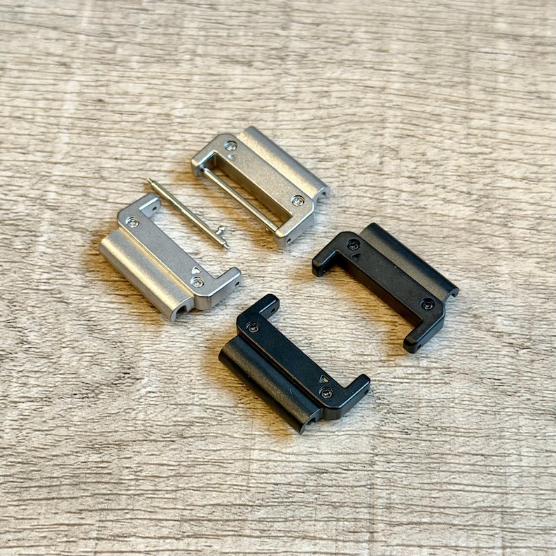 May include: Four watch strap clasps in silver and black. Two silver clasps are rectangular with rounded edges, and one has a silver pin. Two black clasps have a similar design. All are on a light wood grain surface.