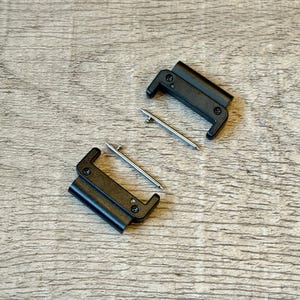May include: Two black watch strap clasps with silver pins, on a light brown wooden surface. The clasps are rectangular with curved ends and small screws. The pins are straight and cylindrical.