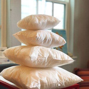 Puede incluir: Un conjunto de cuatro rellenos de almohada blancos, dispuestos en forma de pirámide, sobre una mesa roja. Las almohadas están hechas de un material suave y esponjoso, perfecto para agregar comodidad y soporte a cualquier cama o sofá.